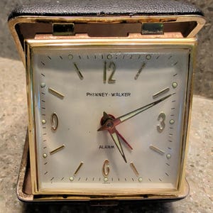 Pode incluir: Um despertador de viagem quadrado Phinney-Walker com moldura de metal dourado e estojo preto com textura de couro. O mostrador creme apresenta marcadores de hora dourados, os números 12, 3, 6 e 9, ponteiros dourados, um ponteiro de alarme vermelho e a palavra ALARM.