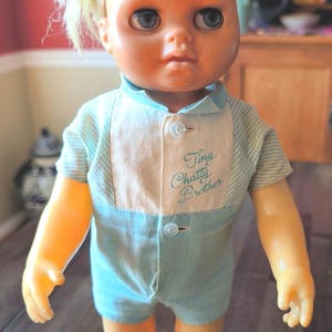 May include: A plastic doll with blonde hair and blue eyes wearing a light blue and white striped romper. The romper features a white chest panel with the embroidered text Tiny Chatty Brother in blue script and two small white buttons down the front.