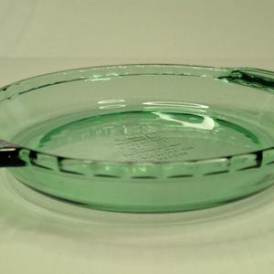 May include: A round, light green glass pie dish with two integrated handles on opposite sides. The rim features a decorative scalloped edge. Embossed text on the bottom interior surface reads Fire-King, Made in U.S.A., and 9 inch pie plate.