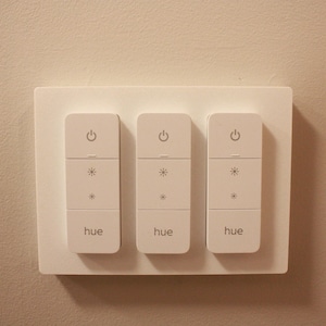 May include: A white rectangular wall-mounted light switch panel featuring three vertical control modules. Each module displays a power icon, two brightness adjustment icons, and the word hue printed in a lowercase sans-serif font.