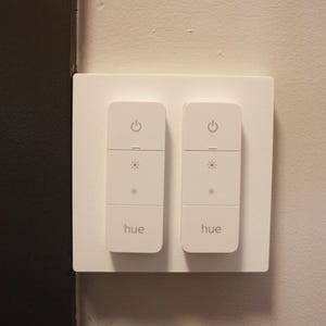 May include: A white dual-gang wall switch plate featuring two vertical smart light controllers. Each controller has a power icon at the top, brightness adjustment icons in the center, and the word hue printed in light gray at the bottom.