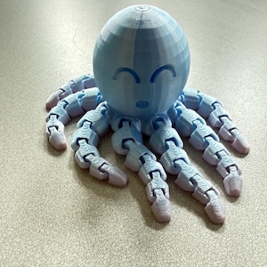 May include: A 3D printed articulated octopus figurine in a light blue color with a gradient shift to a pale pink at the tips of its eight segmented tentacles. The head features a smooth, rounded shape with engraved closed eyes and a small mouth.