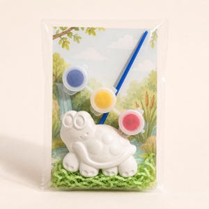 Könnte beinhalten: Eine weiße Schildkrötenfigur aus Keramik, verpackt mit einem blauen Pinsel und drei kleinen runden Farbtöpfen mit blauer, gelber und roter Farbe. Das Set ist auf einer Karte mit einer gedruckten Illustration eines Teichs befestigt.