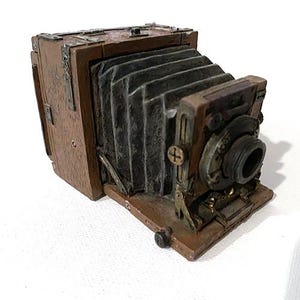 May include: A brown wooden folding camera featuring a dark gray accordion-style bellows, a circular lens assembly, and metallic hardware accents. The device has a rectangular body with visible wood grain texture and a hinged front panel.