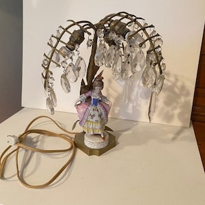 Può includere: Una lampada da tavolo con una statuina centrale in porcellana che raffigura una persona in abiti d'epoca, circondata da una struttura in ottone a forma di salice piangente. Prismi di cristallo trasparenti pendono dai rami e un cavo beige è incluso.