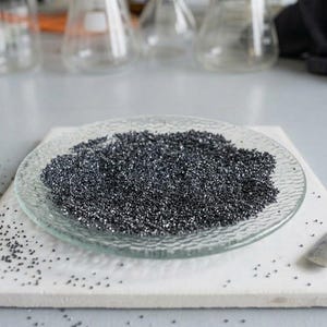 Pode incluir: Um prato circular de vidro transparente com superfície texturizada contém um monte de grânulos finos cinza-escuros com aspeto metálico. Uma pequena espátula de metal prateado repousa sobre a base quadrada branca ao lado do prato, com grânulos espalhados.
