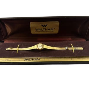 May include: A gold-tone Waltham wristwatch with a textured metal band, presented in a black rectangular display box. The watch face is oval with a white dial. The box interior features the Waltham logo and text including 17 Jewel Custom Design.