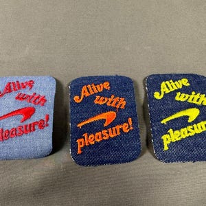 May include: Three rectangular denim patches with rounded corners. The left patch is light blue with red embroidery, the centre is medium blue with orange embroidery, and the right is dark blue with yellow embroidery. All feature the text Alive with pleasure!