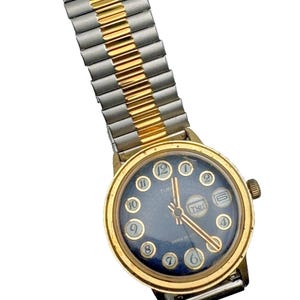 Könnte beinhalten: Eine runde analoge Armbanduhr mit goldfarbener Lünette und dunkelblauem Zifferblatt mit weißen arabischen Ziffern. Die Uhr verfügt über eine Datums- und Wochentagsanzeige auf der Drei-Uhr-Position sowie ein zweifarbiges Metall-Zugband.