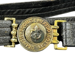 May include: A black textured belt featuring a circular brass buckle. The buckle displays a central fleur-de-lis emblem surrounded by the embossed text SCOUTS DE MEXICO SIEMPRE LISTOS. The belt includes gold-toned metal hardware and two metal eyelets.