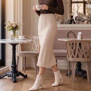 Peut inclure: Une jupe midi couleur crème à taille haute, dotée d'une texture en tricot côtelé vertical et d'une ceinture ajustée. La jupe est associée à un haut à manches longues marron foncé et à des bottines couleur crème à talon bloc et fermeture éclair.