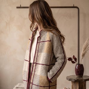 Peut inclure: Un cardigan en tricot à manches longues et ouvert sur le devant, présentant un grand motif à carreaux dans des tons beige, gris et jaune moutarde, rehaussé par un liseré bordeaux foncé le long des bords, du col et des coutures.