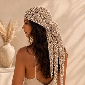 Puede incluir: Un pañuelo de crochet beige con un patrón de punto abierto adornado con pequeñas cuentas blancas. El accesorio para la cabeza se ata en la parte posterior con extremos largos y colgantes que caen, creando un aspecto texturizado de color beige.