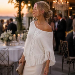 Può includere: Un poncho in maglia bianca con scollo a barchetta, un motivo a trecce verticale al centro e lunghe frange lungo l'orlo inferiore. Il capo è indossato sopra un abito bianco abbinato e completato da una pochette color oro.