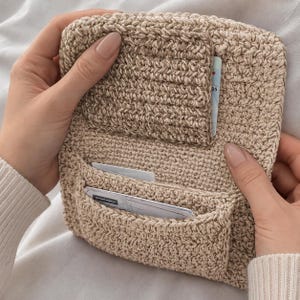 Peut inclure: Un portefeuille en crochet beige doté d'une fermeture à rabat et de plusieurs fentes intérieures. Le portefeuille est fabriqué à partir d'un fil texturé et contient plusieurs cartes en plastique rectangulaires insérées dans les poches.