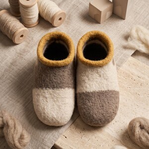 Puede incluir: Un par de zapatillas de lana afieltrada con un diseño de bloques de color en crema y marrón con un borde amarillo mostaza alrededor de la abertura. Las zapatillas se muestran junto a carretes de madera con hilo y fibras de lana sobre tela.