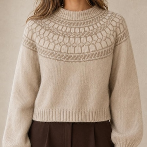 Peut inclure: Un pull en tricot beige avec un col rond et des manches longues à poignets côtelés. La partie supérieure de la poitrine et des épaules présente un motif géométrique détaillé dans une teinte beige plus claire. Le pull est court et duveteux.