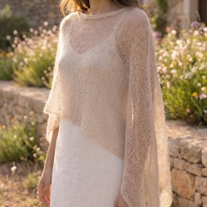 May include: A beige, loose-knit, semi-sheer poncho with a wide boat neckline and a fuzzy, textured finish. The garment features an open, airy weave pattern and drapes over a plain white sleeveless undergarment, creating a layered appearance.
