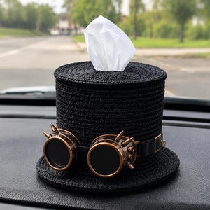Peut inclure: Un porte-mouchoirs cylindrique noir en forme de haut-de-forme, doté d'une surface tissée texturée et d'une paire de lunettes de protection cuivrées à pointes fixées à l'avant. Un mouchoir blanc dépasse de l'ouverture supérieure du chapeau.