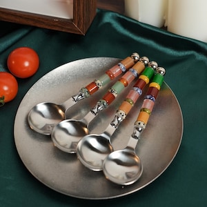 May include: Four silver-toned metal spoons with decorative handles featuring stacked, multi-colored beads in shades of green, orange, red, and blue, along with speckled accents. The spoons are arranged in a fan shape on a round, metallic plate.