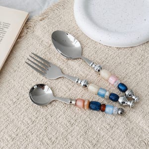 May include: A set of three stainless steel utensils including a large spoon, a fork, and a small spoon. Each handle features a series of colourful, smooth, cylindrical beads in shades of blue, pink, white, and amber, finished with silver metal.