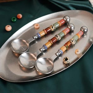 May include: Three silver-toned metal spoons with decorative handles featuring a series of multi-coloured, patterned, and solid-coloured beads. Each handle is topped with a smooth, round silver-toned metal sphere and rests on an oval metal tray.