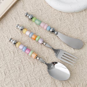 May include: A set of three stainless steel utensils including a butter spreader, a fork, and a spoon. Each handle features a series of smooth, translucent, multi-colored beads in shades of pink, blue, yellow, orange, and green with silver accents.
