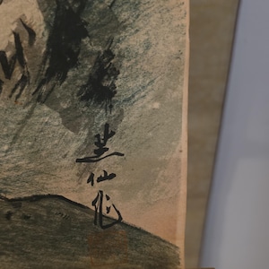 Puede incluir: Un primer plano de una obra de arte en papel texturizado con pinceladas de tinta negra sobre un fondo verde y gris apagado. La pieza incluye caracteres de caligrafía japonesa verticales en tinta negra y un pequeño sello cuadrado rojo.
