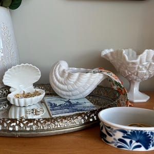Puede incluir: Una colección de recipientes de cerámica blanca que incluye un plato para joyas en forma de concha, un cuenco con forma de caracola con una tela de estampado floral y un cuenco con pie y textura, dispuestos sobre una bandeja redonda de metal plateado.