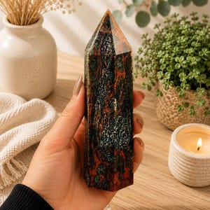 Puede incluir: Una torre de cristal hexagonal pulida con un patr&oacute;n jaspeado en tonos verde bosque profundo, gris carb&oacute;n oscuro y rojo &oacute;xido vibrante. La piedra tiene una superficie lisa y reflectante con una punta facetada y una base plana y estable.