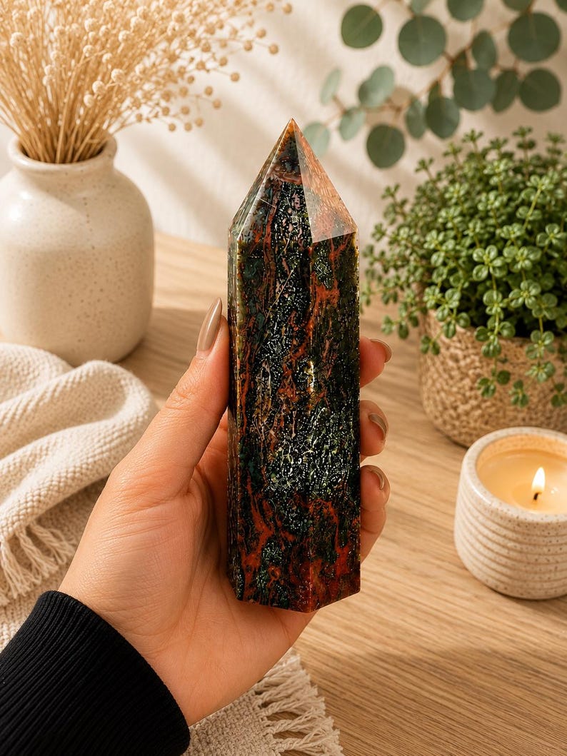 Puede incluir: Una torre de cristal hexagonal pulida con un patr&oacute;n jaspeado en tonos verde bosque profundo, gris carb&oacute;n oscuro y rojo &oacute;xido vibrante. La piedra tiene una superficie lisa y reflectante con una punta facetada y una base plana y estable.