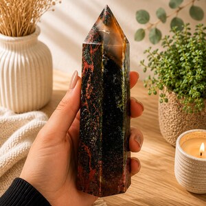 Puede incluir: Una torre de cristal hexagonal pulida con una base oscura y opaca en tonos verde profundo y negro, acentuada por inclusiones de color rojo vibrante y una punta transl&uacute;cida de color &aacute;mbar que capta la luz, mostrando patrones minerales naturales.