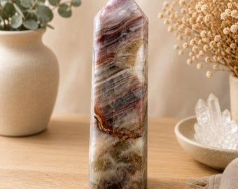 Puede incluir: Una torre de cristal pulido con forma hexagonal y punta superior. La piedra muestra un patrón complejo de colores estratificados que incluyen burdeos intenso, marrón terroso, blanco translúcido y toques de amarillo dorado en la estructura.