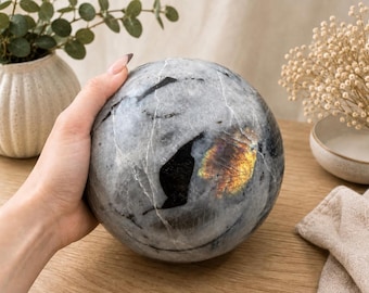 Puede incluir: Una piedra esférica pulida con una base de color gris, vetas blancas prominentes, inclusiones negras profundas y una mancha iridiscente distintiva que refleja tonos amarillos y naranjas sobre su superficie lisa y redondeada.