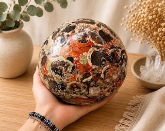 Puede incluir: Una piedra pulida de forma esférica que presenta un patrón complejo y multicolor de tonos rojo intenso, marrón oscuro, negro y verde apagado. La superficie muestra inclusiones intrincadas y formas orgánicas en todo el objeto redondo.