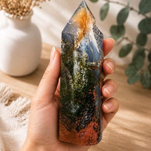 Puede incluir: Una torre de cristal pulido que presenta una compleja mezcla de colores, incluyendo verde bosque profundo, rojo terracota terroso, azul transl&uacute;cido y &aacute;mbar brillante. La piedra tiene una superficie lisa multifac&eacute;tica con inclusiones naturales.