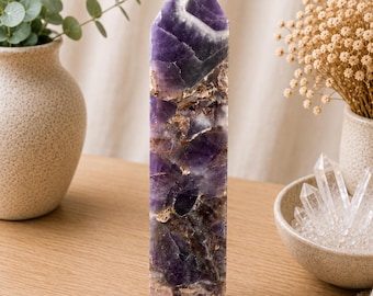 Puede incluir: Una torre de cristal de amatista pulida de forma alta con una punta. La piedra muestra una mezcla de púrpura profundo, violeta translúcido y bandas blancas opacas, con inclusiones minerales naturales de color marrón oscuro y bronce visibles.