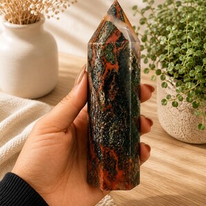 Puede incluir: Una torre de cristal hexagonal pulida que presenta un patr&oacute;n complejo de inclusiones en verde bosque profundo, marr&oacute;n tierra y rojo &oacute;xido vibrante. La piedra tiene una superficie lisa y reflectante con una punta superior y una base plana.