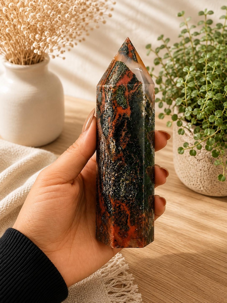 Puede incluir: Una torre de cristal hexagonal pulida que presenta un patr&oacute;n complejo de inclusiones en verde bosque profundo, marr&oacute;n tierra y rojo &oacute;xido vibrante. La piedra tiene una superficie lisa y reflectante con una punta superior y una base plana.