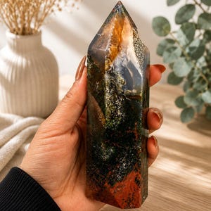 Puede incluir: Una torre de cristal pulido que presenta una mezcla de tonos verde bosque profundo, marr&oacute;n terroso y &aacute;mbar transl&uacute;cido. La piedra tiene una superficie lisa y multifac&eacute;tica con inclusiones naturales que crean un patr&oacute;n similar a un paisaje.