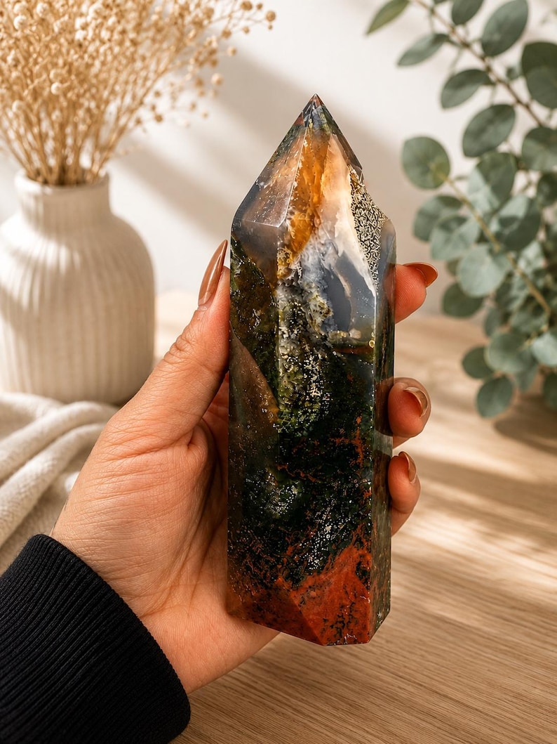Puede incluir: Una torre de cristal pulido que presenta una mezcla de tonos verde bosque profundo, marr&oacute;n terroso y &aacute;mbar transl&uacute;cido. La piedra tiene una superficie lisa y multifac&eacute;tica con inclusiones naturales que crean un patr&oacute;n similar a un paisaje.