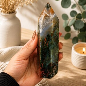 Puede incluir: Una torre de cristal de &aacute;gata musgo pulida con forma de prisma hexagonal y punta superior. La piedra muestra una mezcla compleja de inclusiones en verde bosque profundo, marr&oacute;n terroso y blanco transl&uacute;cido con patrones minerales.
