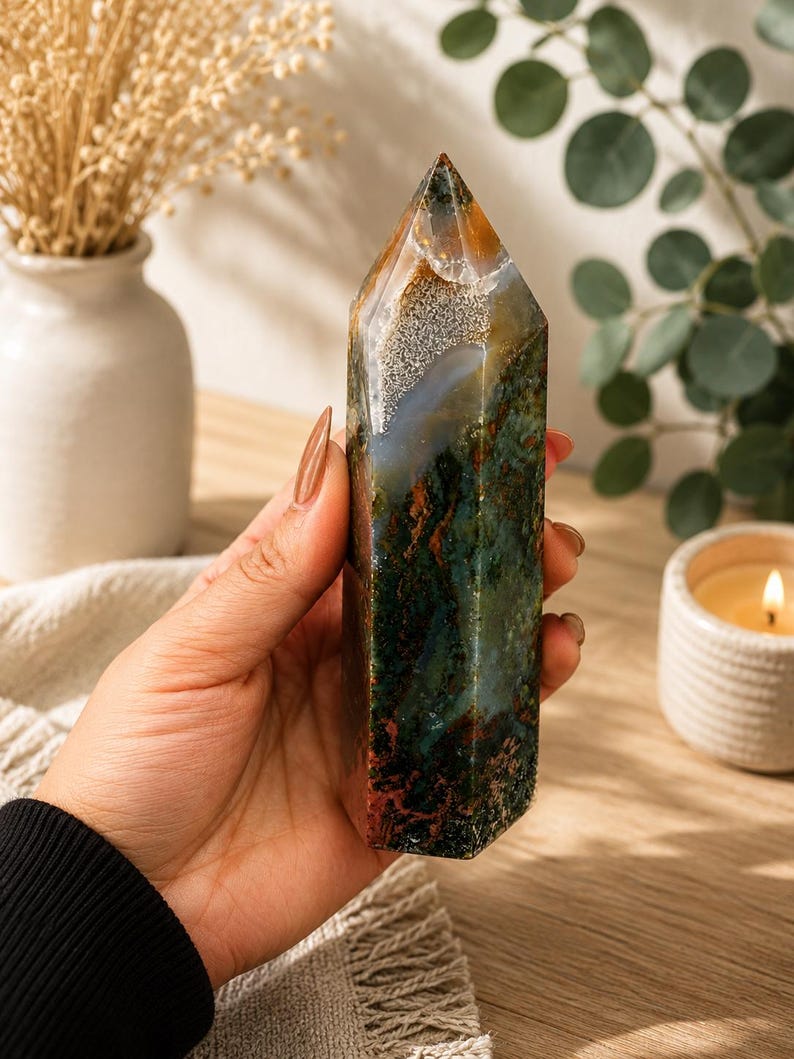 Puede incluir: Una torre de cristal de &aacute;gata musgo pulida con forma de prisma hexagonal y punta superior. La piedra muestra una mezcla compleja de inclusiones en verde bosque profundo, marr&oacute;n terroso y blanco transl&uacute;cido con patrones minerales.