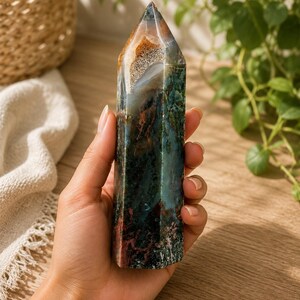 Puede incluir: Una torre de cristal hexagonal pulida que presenta una mezcla compleja de inclusiones minerales en verde bosque profundo, marr&oacute;n tierra y blanco transl&uacute;cido. La piedra tiene una superficie lisa y multifacetada con una punta y un patr&oacute;n natural.