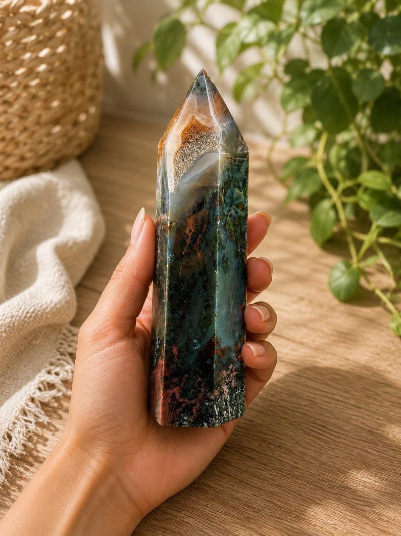 Puede incluir: Una torre de cristal hexagonal pulida que presenta una mezcla compleja de inclusiones minerales en verde bosque profundo, marr&oacute;n tierra y blanco transl&uacute;cido. La piedra tiene una superficie lisa y multifacetada con una punta y un patr&oacute;n natural.