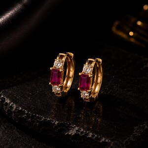 May include: A pair of gold hoop earrings, each featuring a central rectangular-cut deep red gemstone flanked by two small round-cut clear white stones. The earrings have a polished gold finish and a hinged closure mechanism for secure wear.
