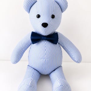 Op de afbeelding: Een knuffelbeer gemaakt van lichtblauwe en witte gestreepte stof. De beer heeft twee ronde zwarte knoopogen, een kleine zwarte neus en een effen donker marineblauwe vlinderdas bij de nek. De ledematen en oren zijn afgerond.
