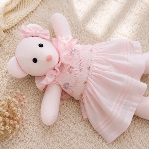 Peut inclure: Un ours en peluche rose clair avec des yeux en perles noires et un nez rose. L'ours porte une robe rose clair assortie avec un corsage smocké, un col à volants et de petites broderies florales. Un petit nœud rose est fixé sur la tête de l'ours.