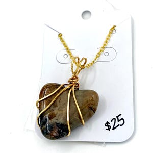 May include: A heart-shaped brown and black stone pendant wrapped in gold-colored wire, suspended from a matching gold-colored metal chain. The necklace is displayed on a white card with the text $25 printed in black ink on the lower right side.