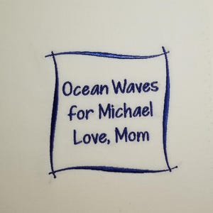 May include: A white fabric with blue embroidery that reads "Ocean Waves for Michael Love, Mom" inside a hand-drawn square.